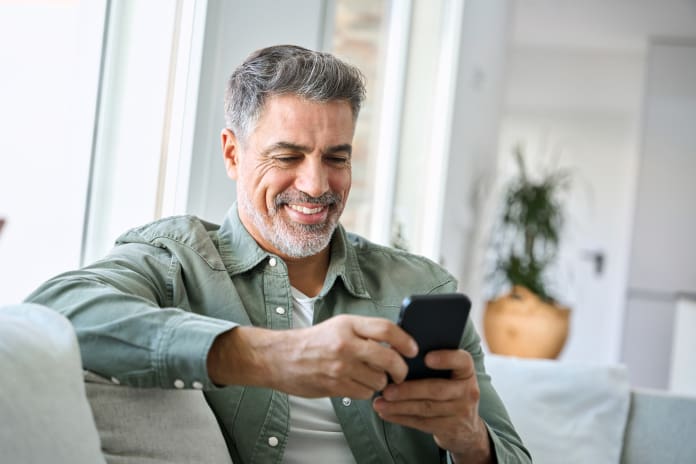 Smiling mature middle aged man using mobile phone sitting at home on couch.
